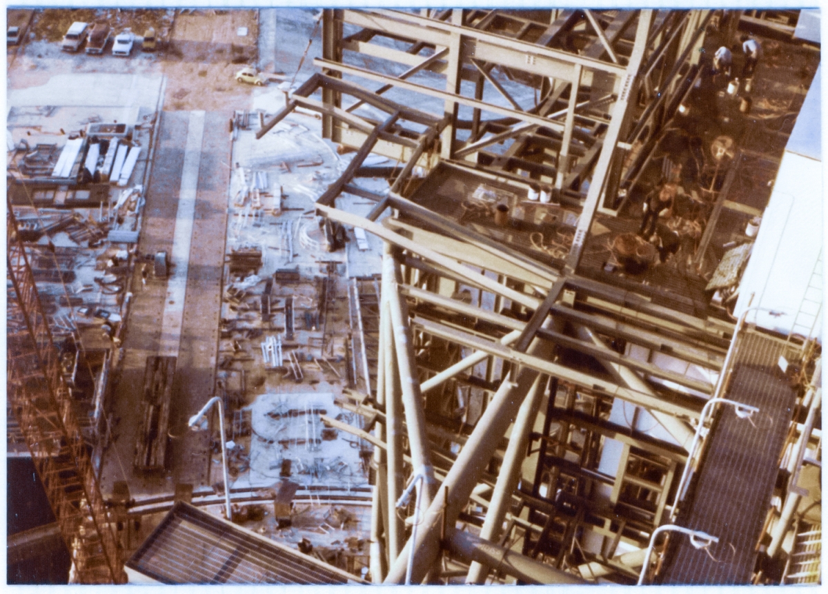 Union Ironworkers from Local 808, assembling the steel skeleton of the Rotating Service Structure at Space Shuttle Launch Complex 39-B, Kennedy Space Center, Florida. On the ground, in the distance, a multitude of steel elements lay on either side of one trackway of the crawlerway, waiting to be lifted into place on the growing structure.